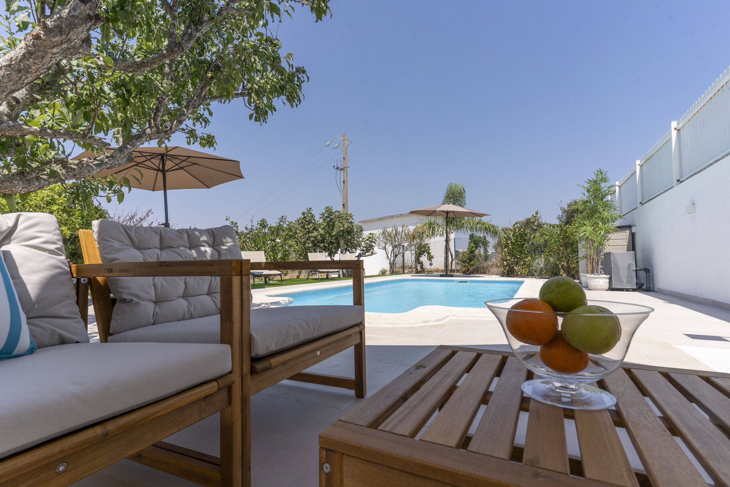 Un espace aménagé au bord de la piscine, avec un fauteuil de relaxation, une table en bois sur laquelle sont disposés des fruits et des parasols.