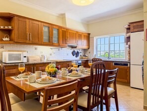 A kitchen with wooden cabinets, a microwave, and a dining table set with plates, glasses, and a fruit bowl. - #373 Charming 4-Bedroom Villa with a Spacious Garden & BBQ (Albufeira)