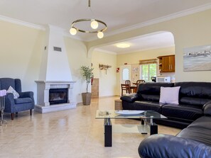 A living room with a fireplace, a black leather sofa, a glass coffee table, and a dining area in the background. - #373 Charming 4-Bedroom Villa with a Spacious Garden & BBQ (Albufeira)