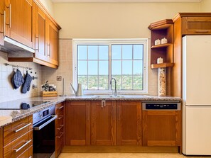 A kitchen with wooden cabinets, a built-in oven, a dishwasher, and a window with a view. - #373 Charming 4-Bedroom Villa with a Spacious Garden & BBQ (Albufeira)