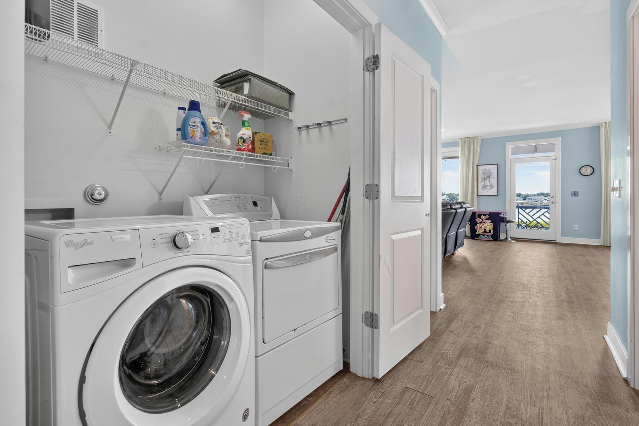 A laundry room with a front-loading washer and dryer, shelves with cleaning supplies, and a view of the living area through an open door.