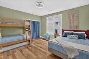 Een kamer met stapelbedden, een enkel bed, een plafondventilator, een raam met jaloezieën, een macramé wanddecoratie en een kleine tafel met spullen.