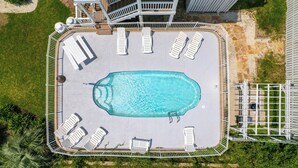 Eine Luftaufnahme eines rechteckigen Schwimmbeckens mit einer umlaufenden Holzterrasse, umgeben von einem Zaun, neben einem Gebäude mit einer Treppe.
