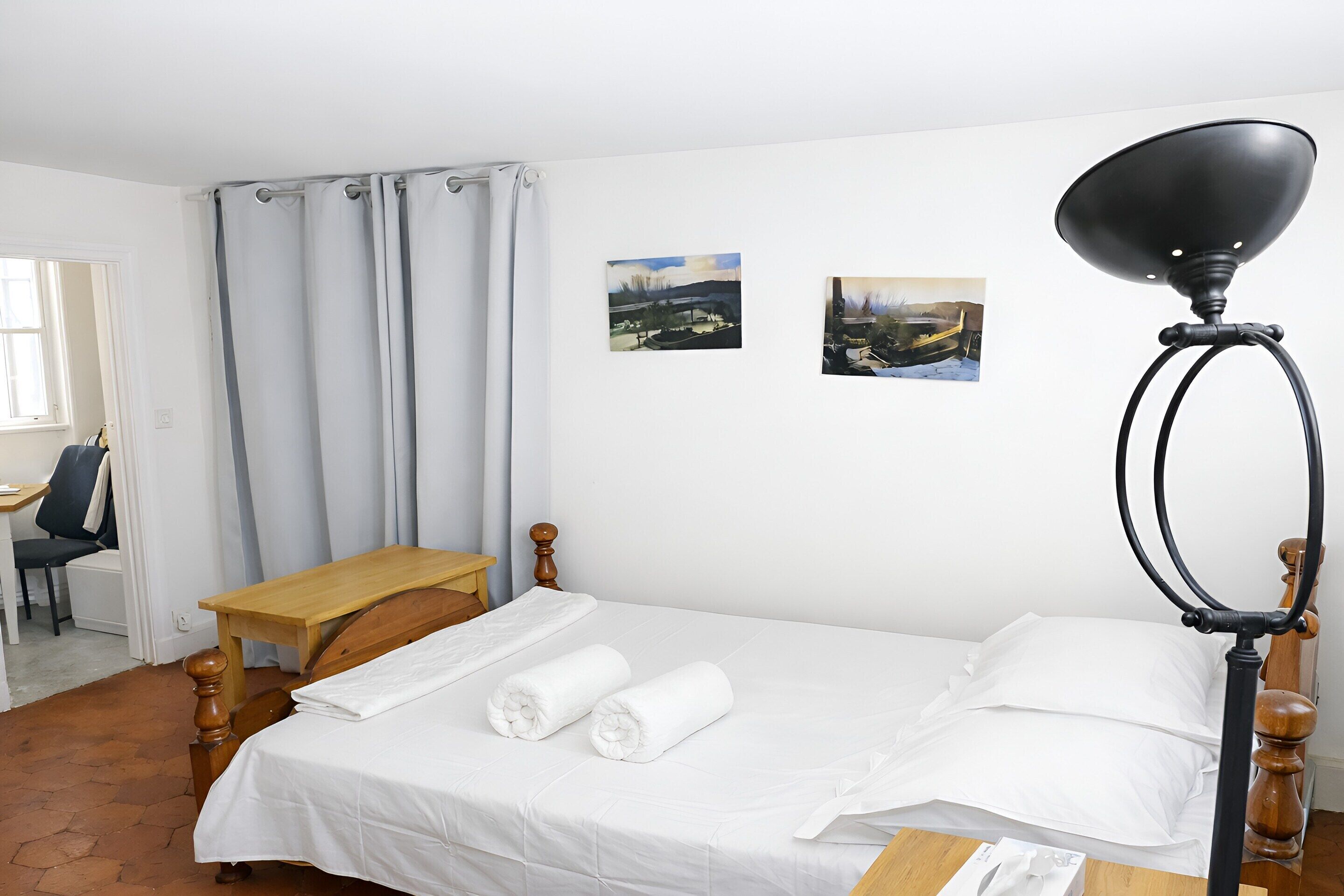 Una cama bien hecha con ropa blanca, una mesita de noche de madera, un escritorio y una silla en una habitación con paredes blancas y una ventana con cortinas.
