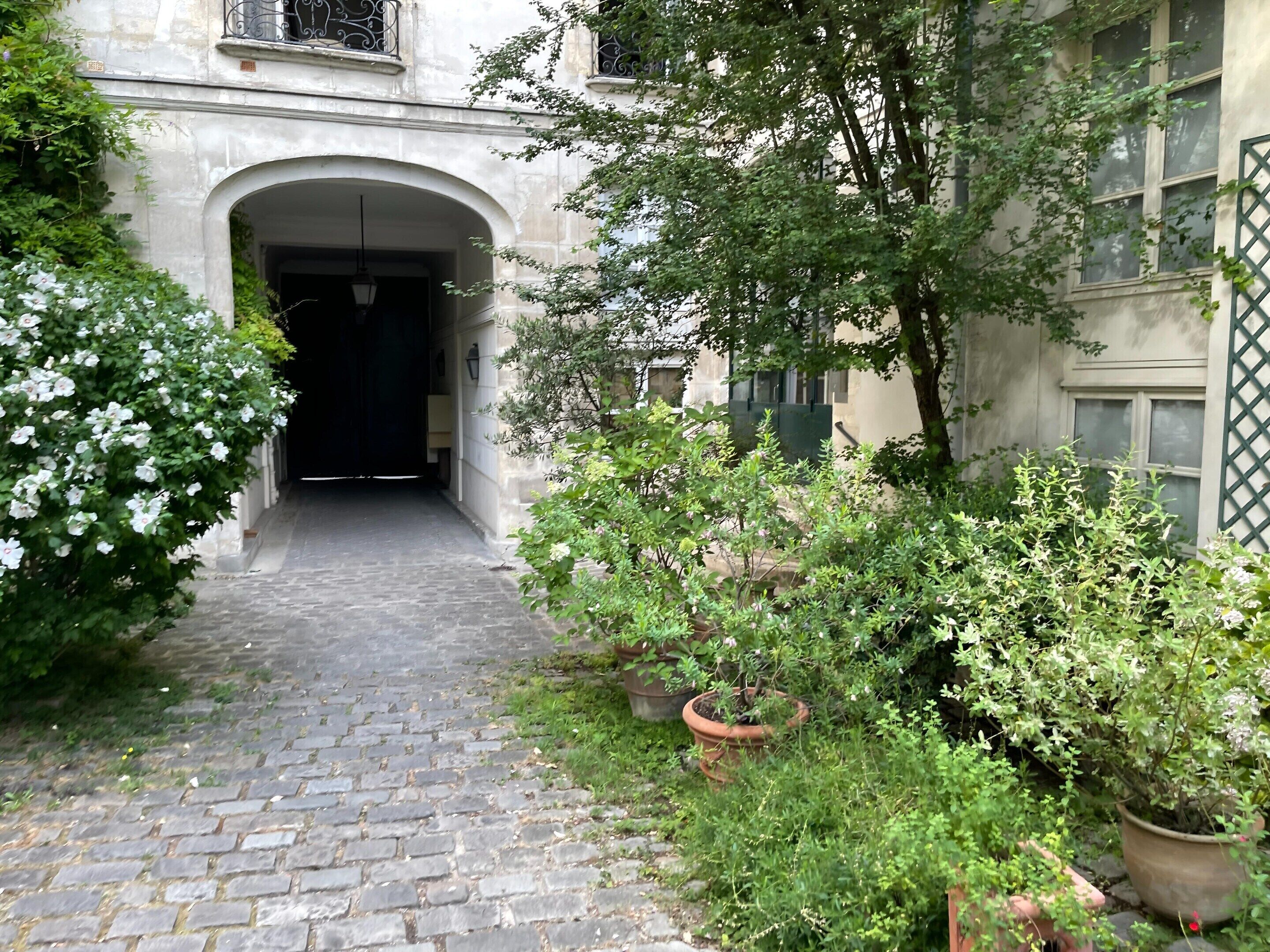 Un chemin pavé de pierres menant à l’entrée d’un bâtiment, agrémenté d’un portail en fer forgé, le tout entouré de verdure et de plantes en pot.