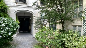 Un chemin pavé de pierres menant à l’entrée d’un bâtiment, agrémenté d’un portail en fer forgé, le tout entouré de verdure et de plantes en pot.