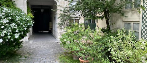 Een geplaveid pad dat leidt naar de ingang van een gebouw met een ijzeren hek, omgeven door groen en plantenpotten.