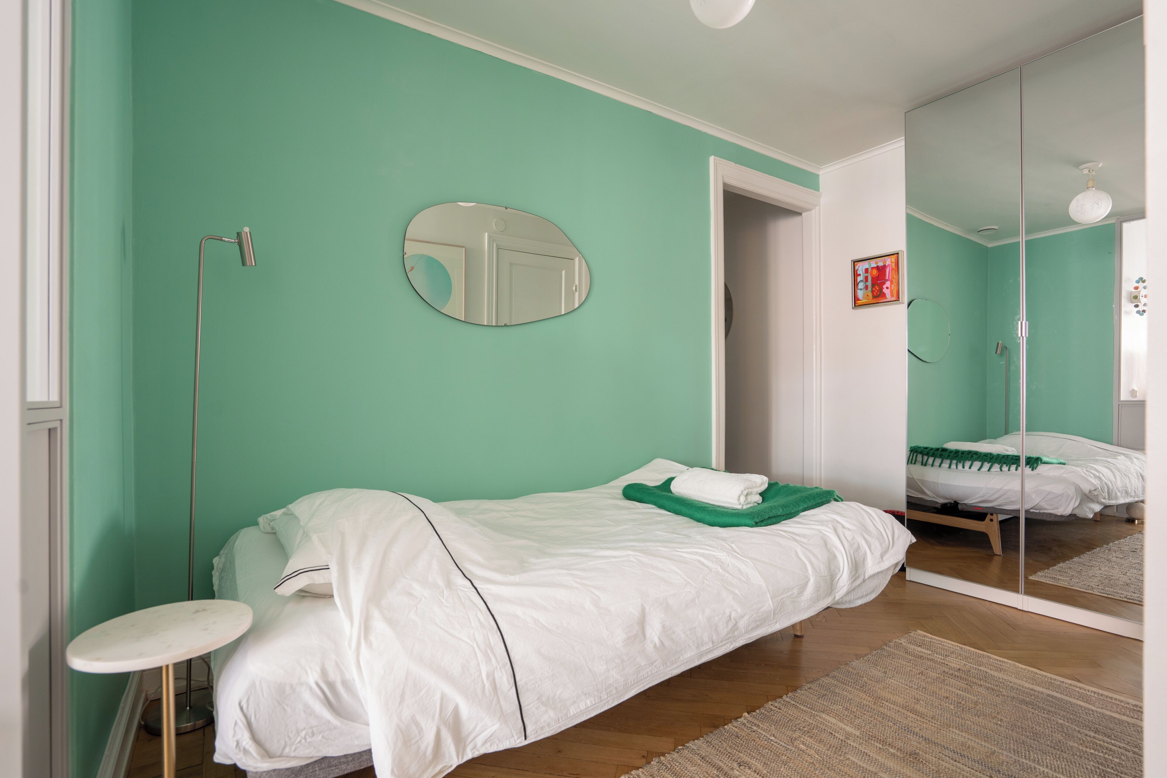 A bedroom with a bed, a round mirror, a bedside table, and a floor lamp.