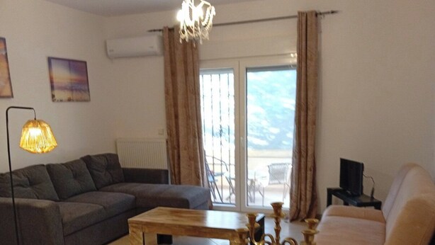 A living room with a sofa, a dining table with chairs, a chandelier, and a view through a glass door. - Cozy1-bedroom apartment, two sofa bed in enchanting peraia-neoi epivates with AC (Thessaloniki)