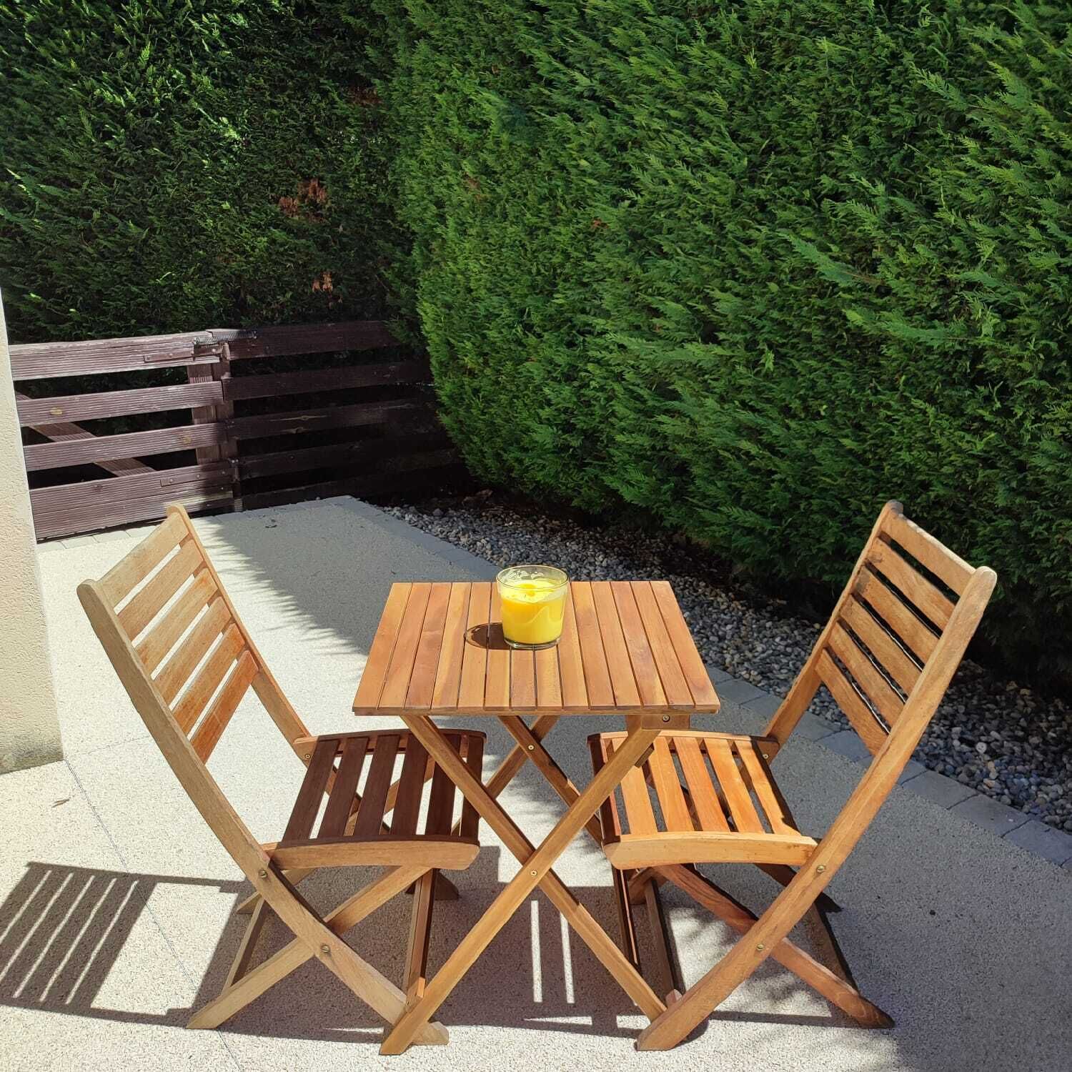 Een houten tafel met twee stoelen op een betonnen terras, met een haag op de achtergrond.