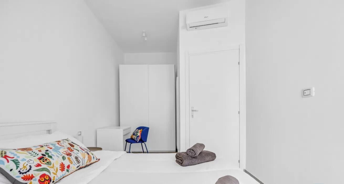 A minimalist hotel room with a bed, a chair, a wardrobe, and a rolled towel on the floor.