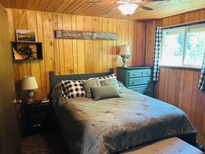 A bedroom with a wooden paneled wall, a bed with a quilt, a nightstand with a lamp, a dresser, and a window with curtains. - Family Friendly, Cozy 3-bedroom cabin in amazing Cascade with AC and Wood stove. (Cascade)