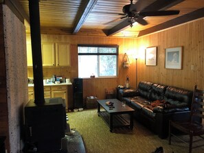A cozy living room with a leather sofa, a coffee table, and a fireplace. There is a kitchen area with wooden cabinets and a window. - Family Friendly, Cozy 3-bedroom cabin in amazing Cascade with AC and Wood stove. (Cascade)