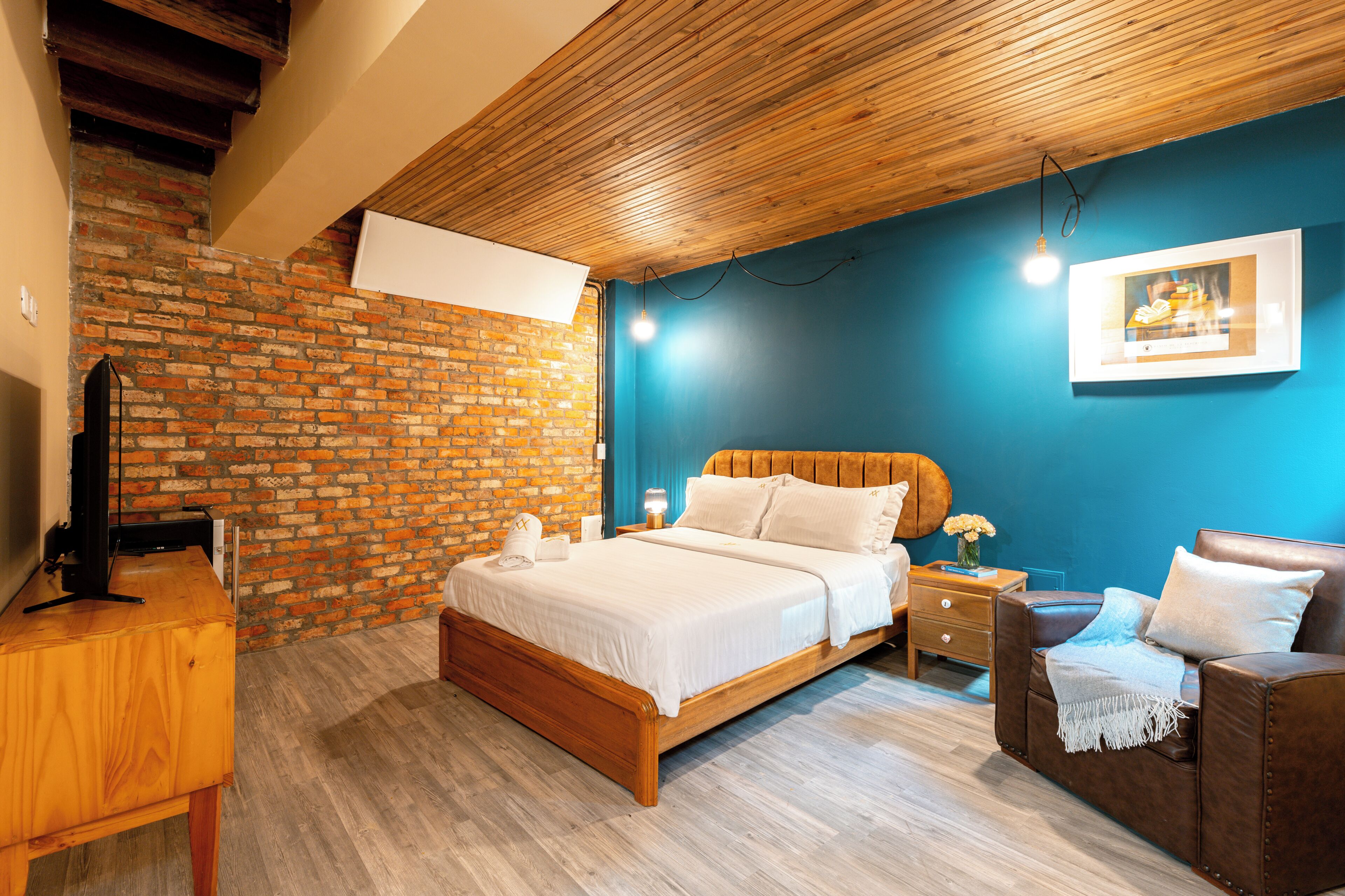 A bedroom with a wooden ceiling, a brick wall, a blue accent wall, a bed with white bedding, a wooden nightstand, a brown leather chair, and a framed picture on the wall.