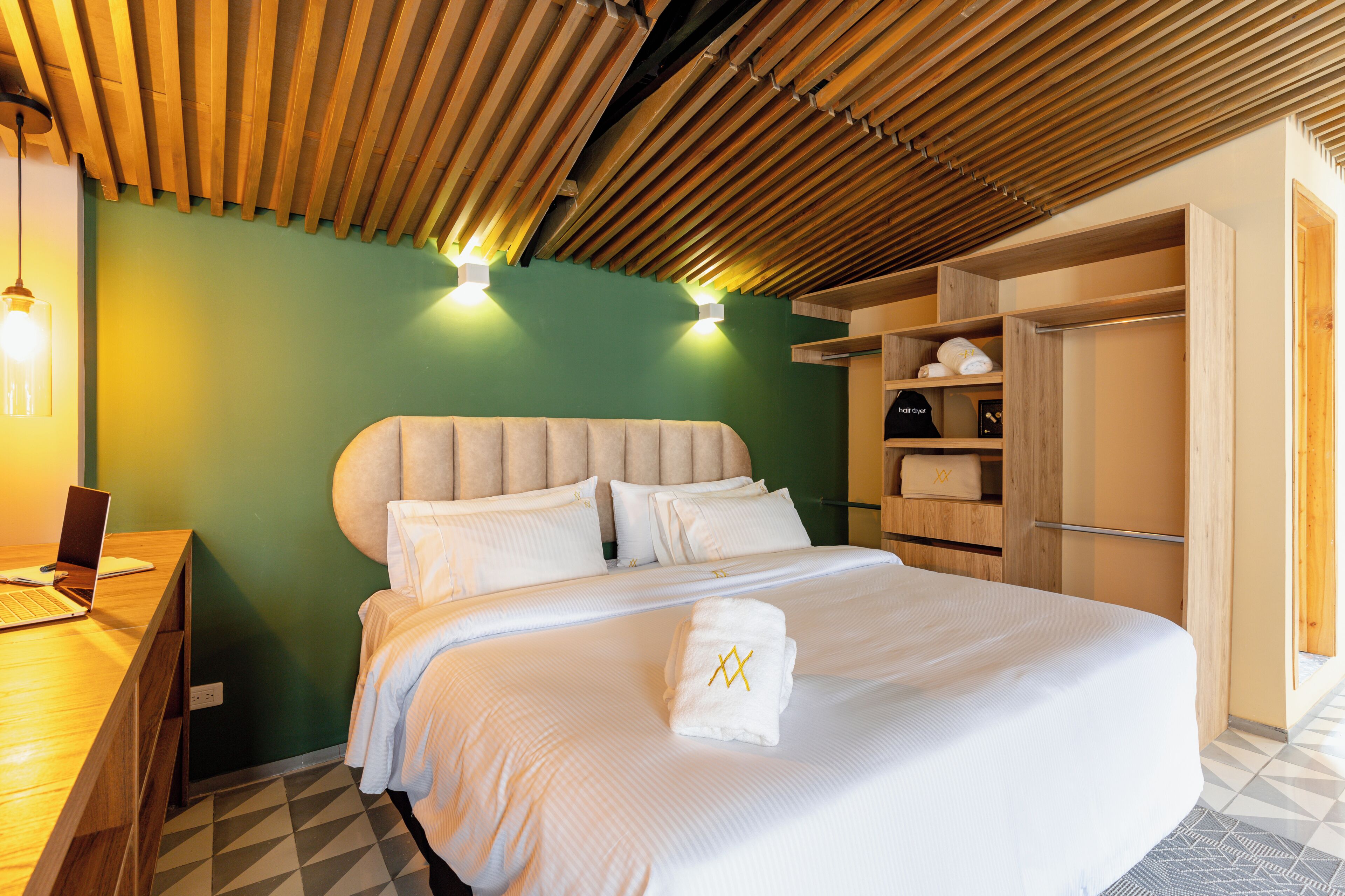 A hotel room with a bed, a desk with a laptop, a wardrobe, and a green wall.