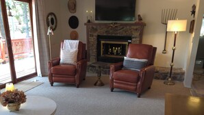 Un salon comprenant deux fauteuils en cuir, une table basse ronde, une cheminée avec un manteau en pierre et un ventilateur de plafond.