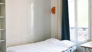 Ein Einzelbett mit weißer Decke, ein kleiner Tisch und ein Fenster mit Vorhängen.