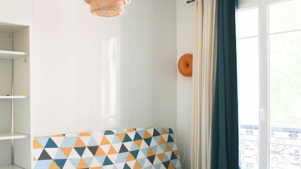 A room with a patterned sofa, a hanging light fixture, a window with curtains, and a balcony with a railing.
