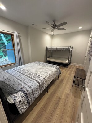 A compact bedroom with a bunk bed, a single bed with patterned bedding, a ceiling fan, and a window with curtains.