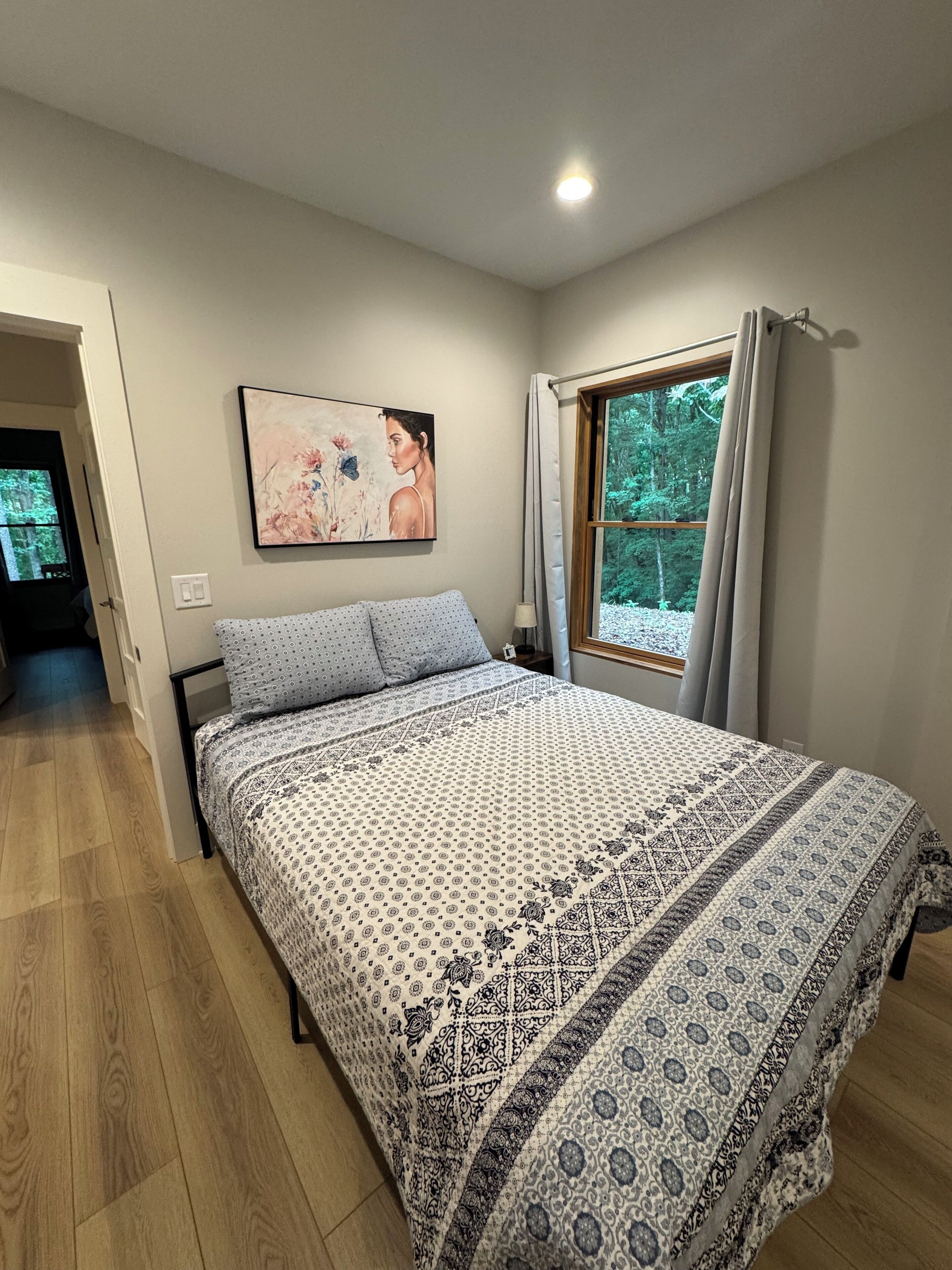 A bedroom with a bed, a painting, a window with curtains, and a door leading to another room.