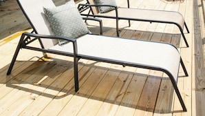 Eine Holzterrasse mit Liegestühlen und einem Tisch, ein überdachter Bereich mit Geländer und ein Holzbau im Hintergrund.