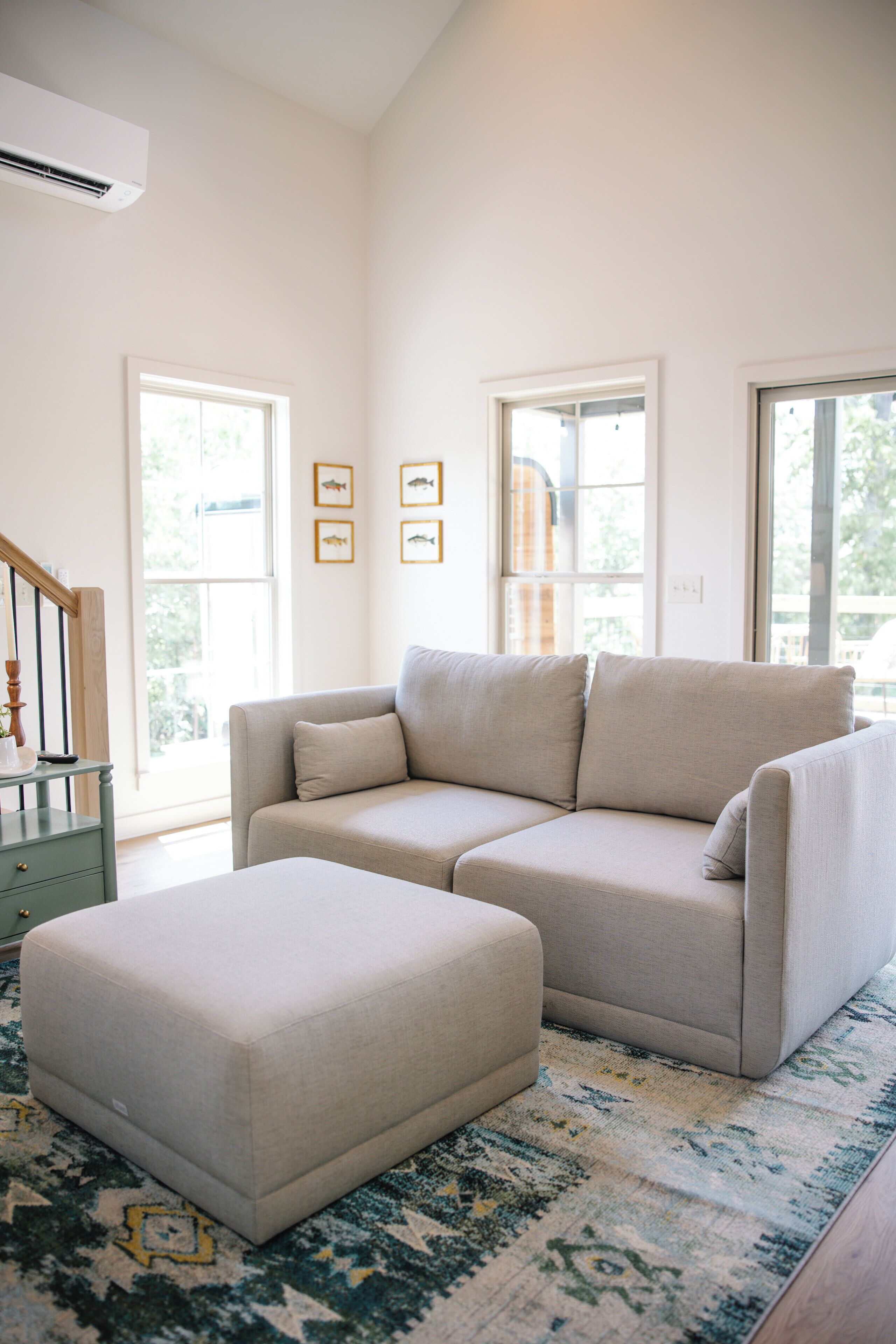 A modern living room with a sectional sofa, a matching ottoman, and a patterned rug. There are large windows providing natural light and a view of the outdoors.