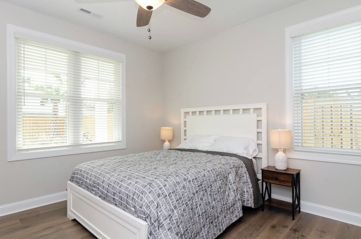 A bedroom with a bed, two bedside tables with lamps, a ceiling fan, and large windows with blinds.