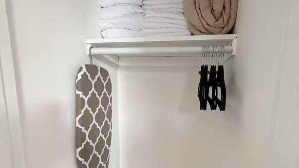 Um espaço de armazenamento com cobertores brancos e bege dobrados, um pendurar de parede estampado e uma estante com artigos pretos.