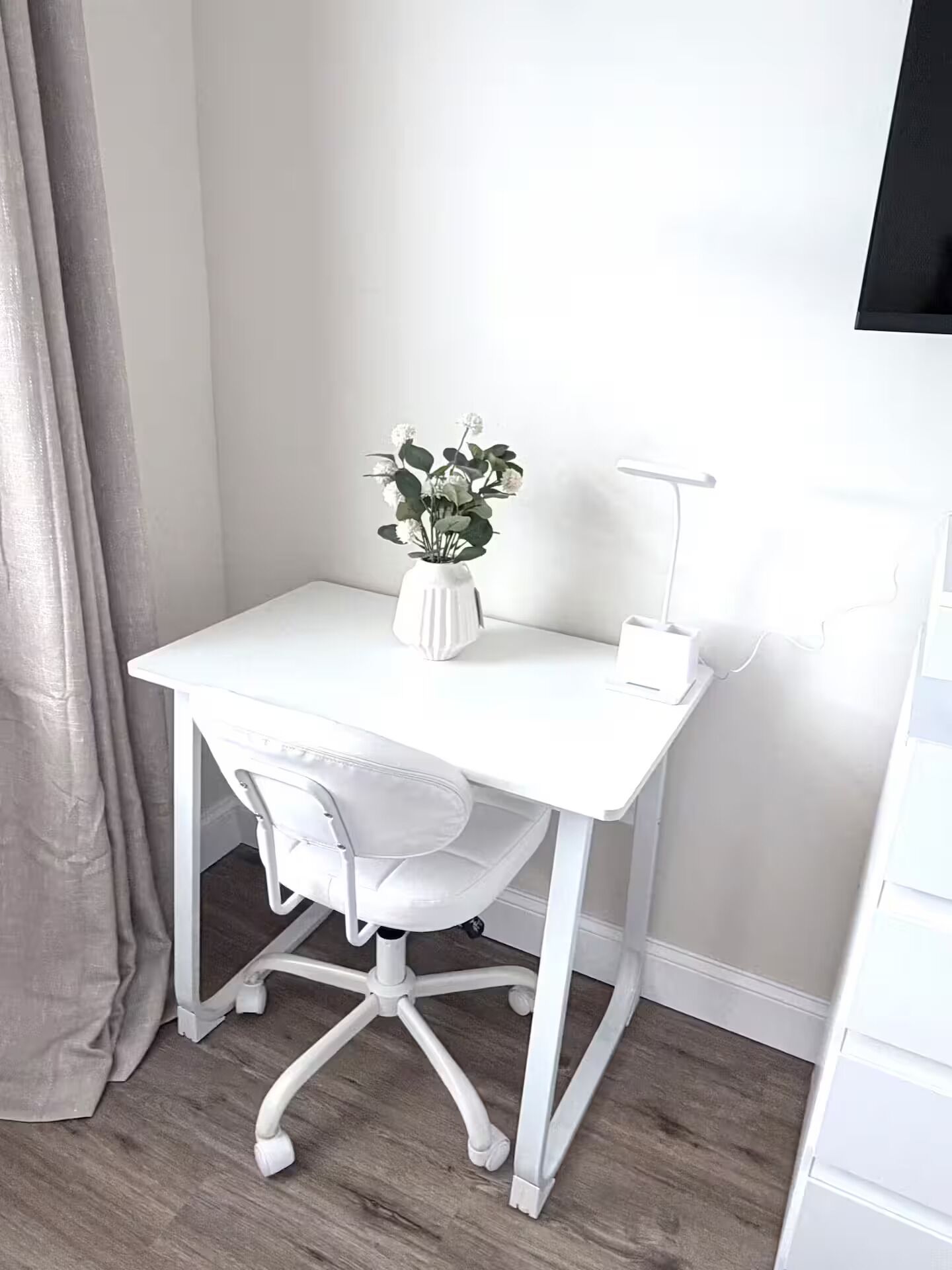 A white desk with a chair, a white vase with flowers, and a desk lamp.