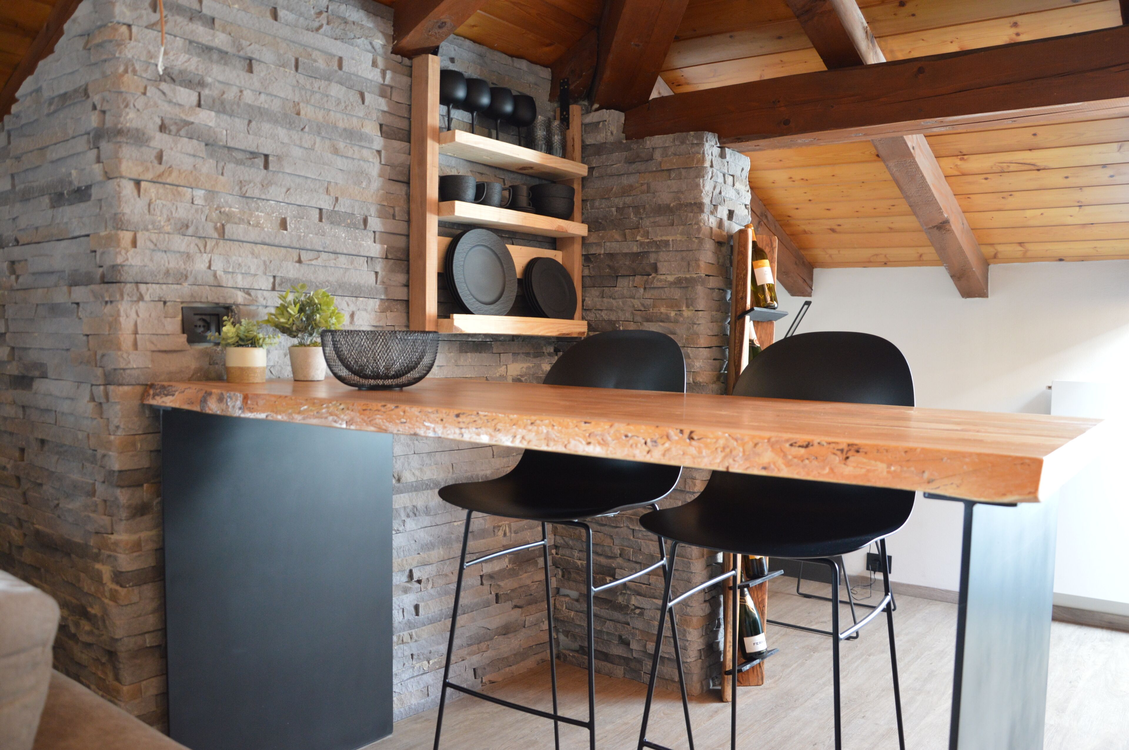 Een keuken met een stenen muur, houten balken, een bargedeelte en zwarte stoelen.