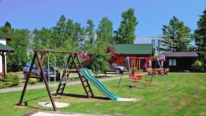 Leikkikenttä, jossa on keinut ja liukumäki, ja jonka ympärillä on nurmikkoalue ja puita.
