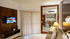 A modern living room with a flat-screen TV, a sofa, a coffee table, and a kitchen area visible through an open door. - SHB Elegante apto com vista mar  (Santos)