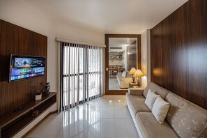 A modern living room with a flat-screen TV mounted on a wooden wall, a sofa with cushions, a lamp on a side table, and a sliding glass door leading to a kitchen area. - SHB Elegante apto com vista mar  (Santos)