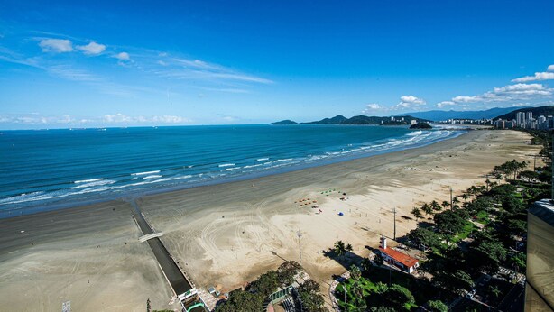 A coastal beach with a clear sky, a sandy shore, and a distant cityscape. - SHB Elegante apto com vista mar  (Santos)