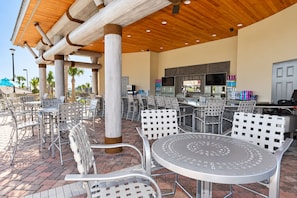 An outdoor patio area with metal chairs and tables, a wooden ceiling structure, and a bar area with a television and various items on shelves. - 9BR Resort Home for Large Groups with Private Pool Near Disney (Four Corners)