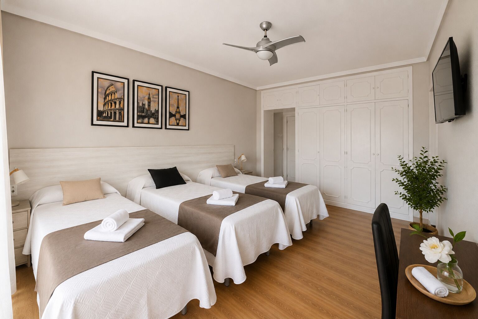 Et hotelværelse med tre senge, en ventilator, et fjernsyn og et lille bord med en plante.
