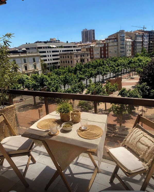 A balcony with a table set for two, surrounded by wicker chairs, with a view of a cityscape and trees.