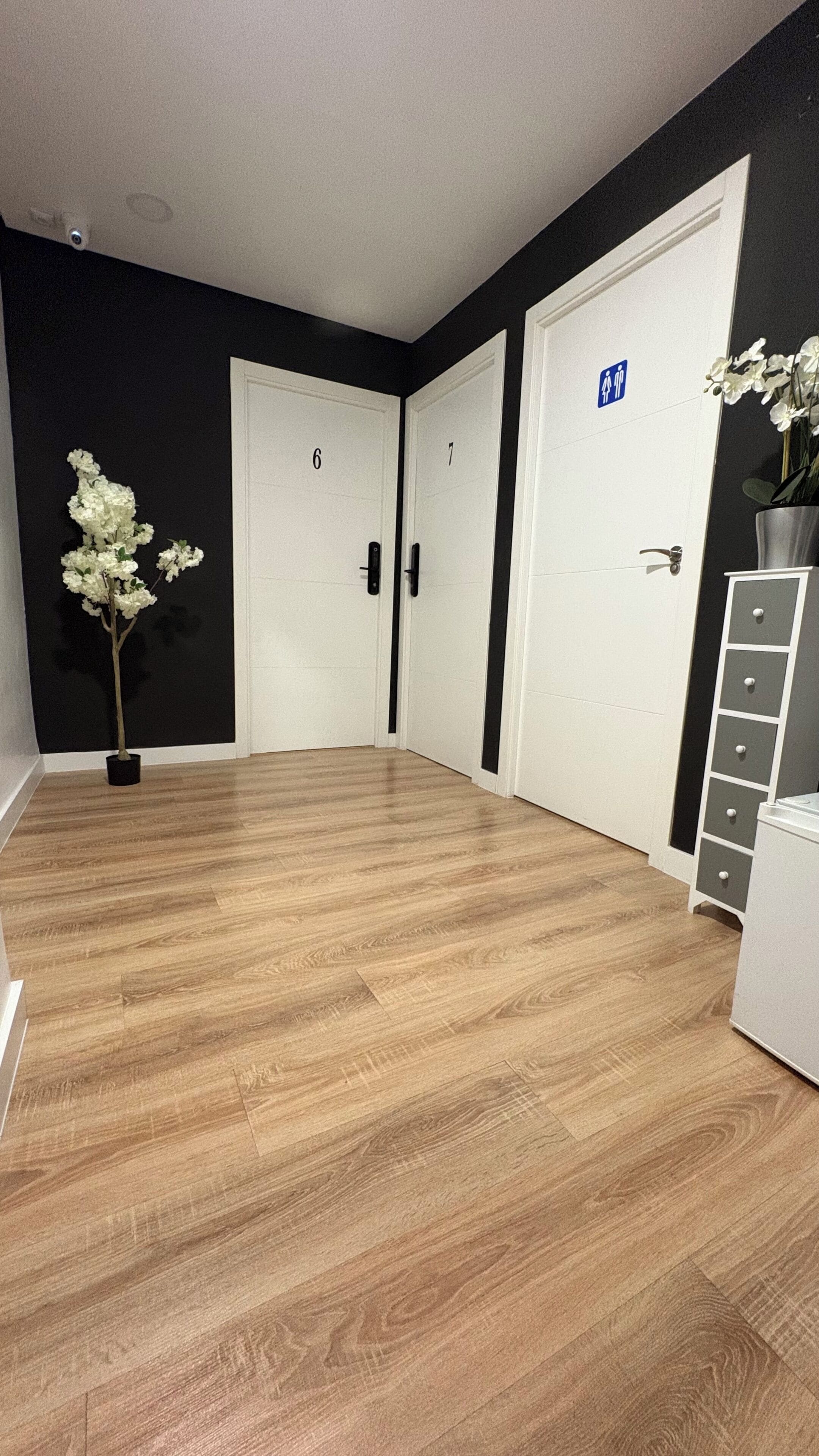 Un pasillo con piso de madera, puertas blancas numeradas 6 y 7, y un letrero azul que indica el baño de hombres.