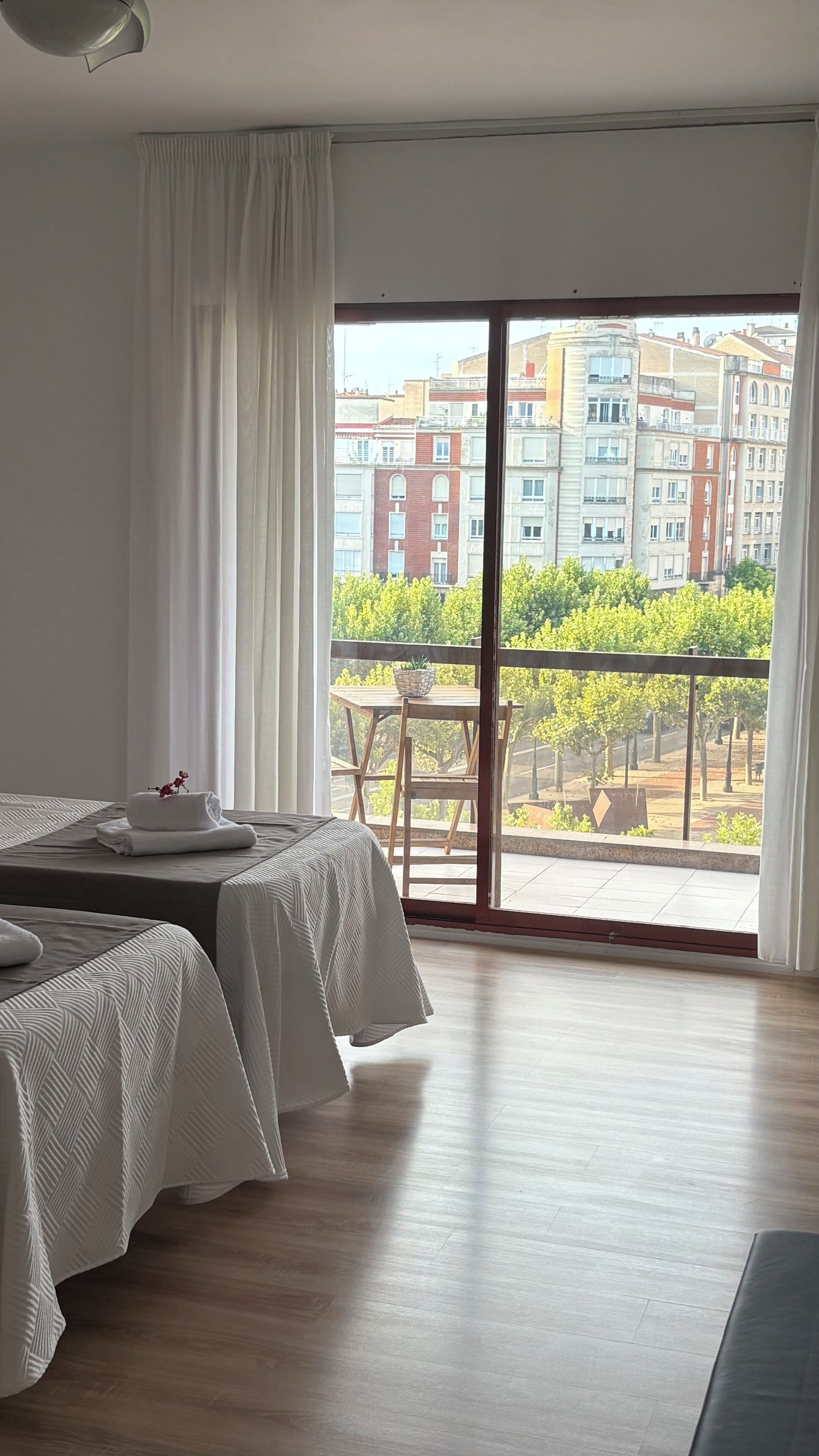 Une chambre d’hôtel avec deux lits, un balcon doté d’une table et de chaises, et une vue sur des immeubles et des arbres.