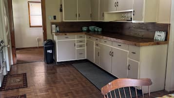 Ein Zimmer mit Holzboden, einem Bett mit floralem Überwurf, einem Holzstuhl, einer Küche mit weißen Schränken und einem Fenster mit Vorhängen.