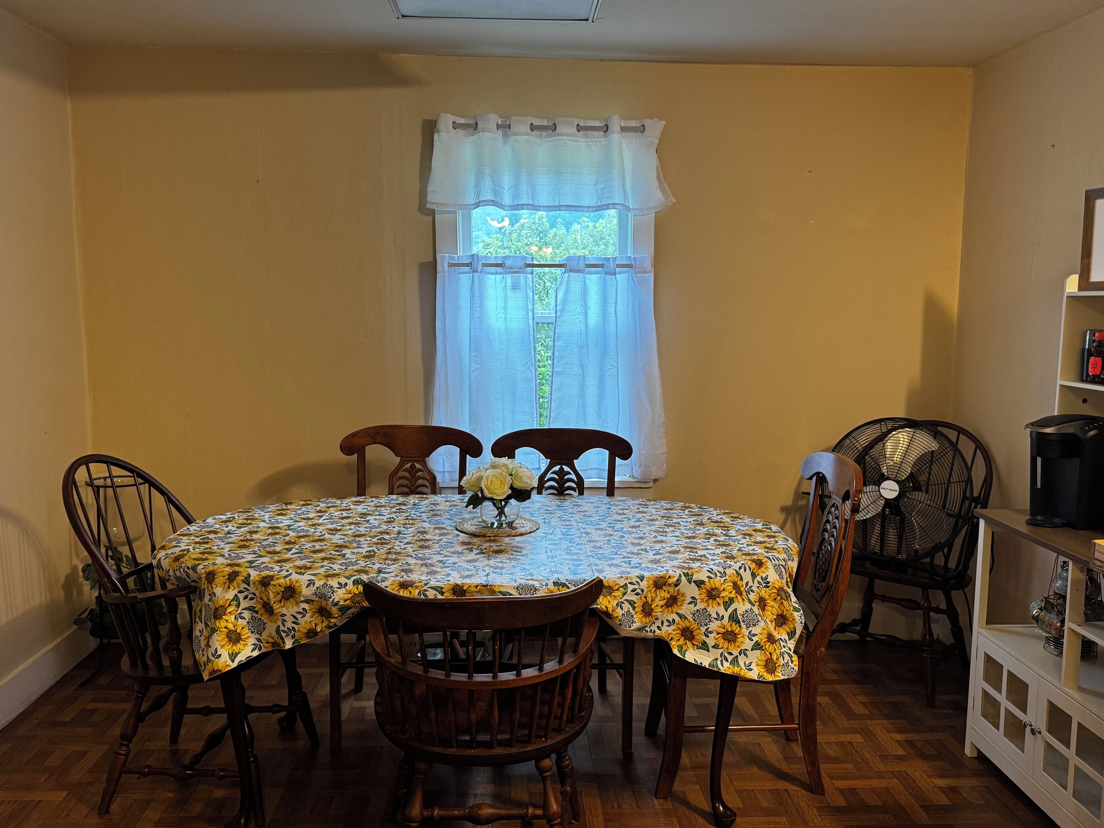 A dining area with a round table covered in a sunflower-patterned tablecloth, surrounded by wooden chairs.