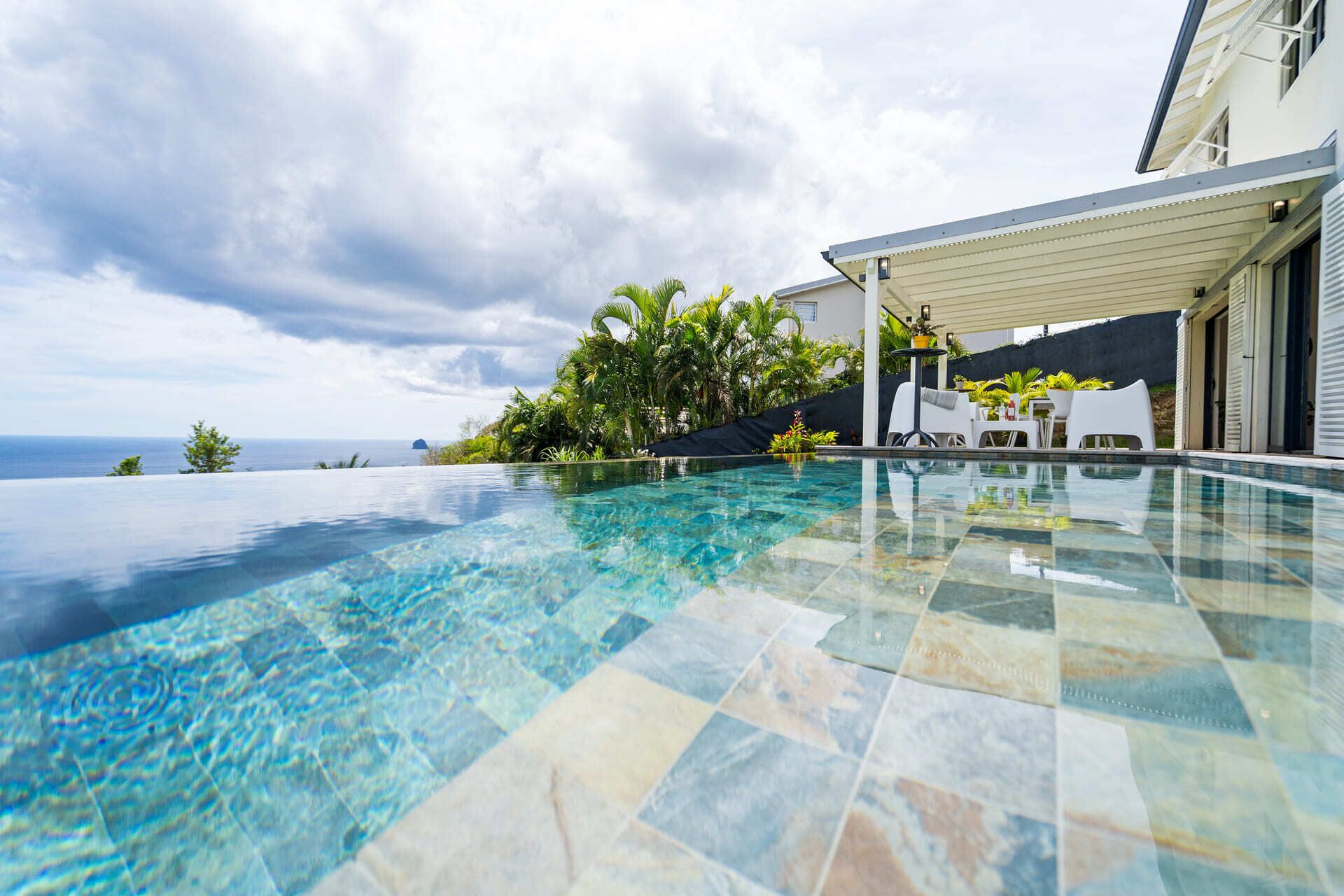 An infinity pool with a clear view of the ocean, a covered outdoor seating area, and lush tropical plants.