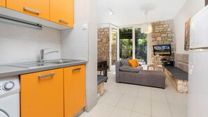 A modern kitchen with orange cabinets, a built-in oven, and a dining area with a grey sofa and a TV. - Garden Apartment Orange I Porec Old Town (Porec)