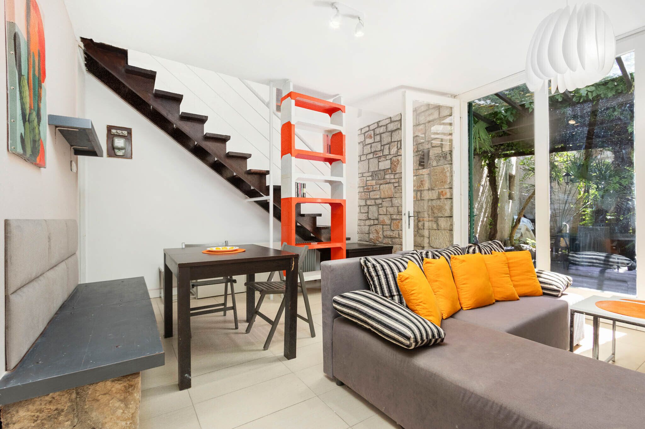 A modern living space with a staircase, a sofa with striped cushions, a dining table, and a stone accent wall.