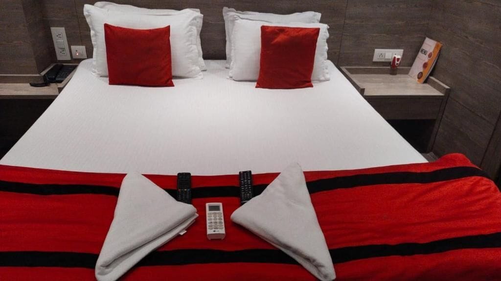 A hotel room with a bed, red and white pillows, a red and black striped blanket, and a remote control.