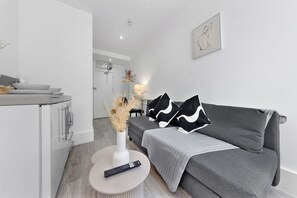 A modern living room with a grey sofa, a white vase with dried plants, and a remote control on a round table. - West London Stay Studio (London)