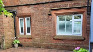 Une maison en briques avec deux fenêtres, un petit jardin agrémenté de plantes en pot et une cour dallée de pierres.