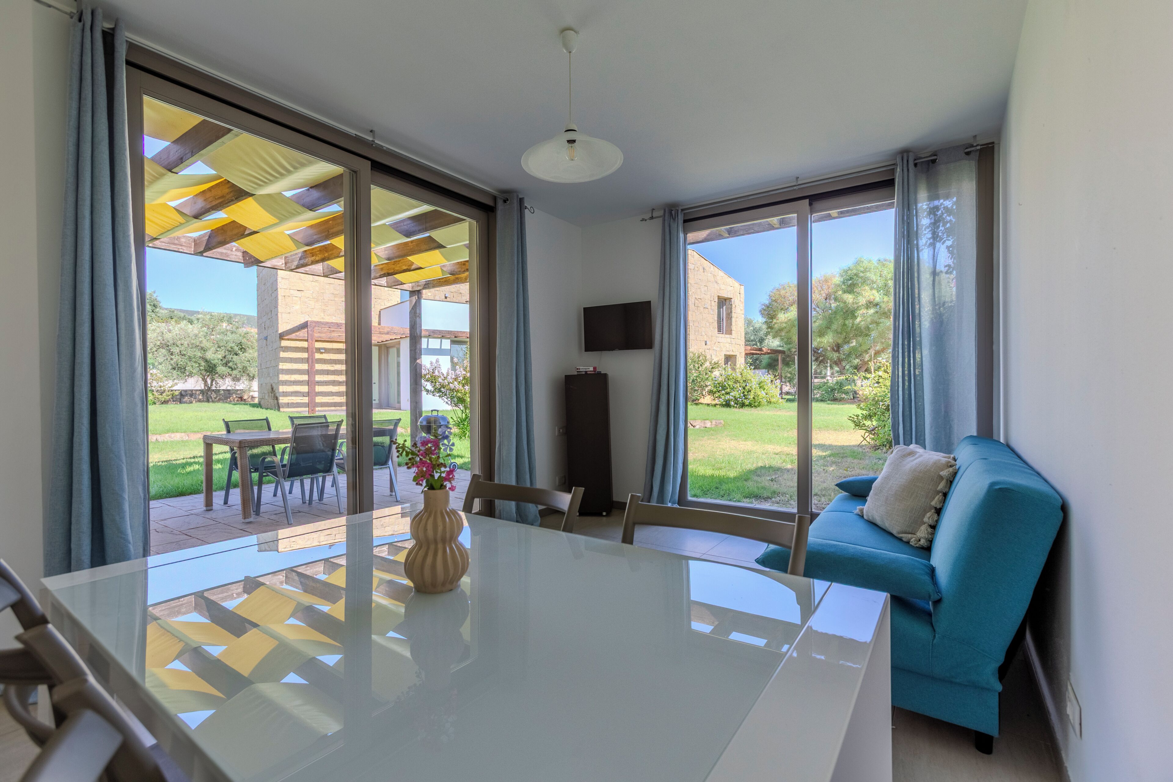 A modern living room with a glass table, a blue sofa, and a TV mounted on the wall.