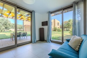Ein Zimmer mit einem blauen Sofa, einem an der Wand montierten Fernseher und einer Glastür, die auf eine Außenpatio mit Tisch und Stühlen führt.