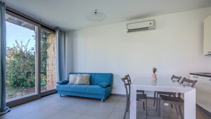 A modern living room with a blue sofa, a dining table with chairs, a large window with curtains, and an air conditioning unit on the wall. - Golden Stone Villa: Pool & Relax (Masainas)